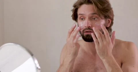 Young shirtless man looking at small mirror and applying facial cream. Bearded Stock Footage 142529122