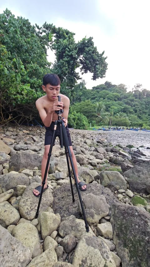 Young Shirtless Man Setting Up Camera Tripod On A Rocky Beach Stock Footage 339829208