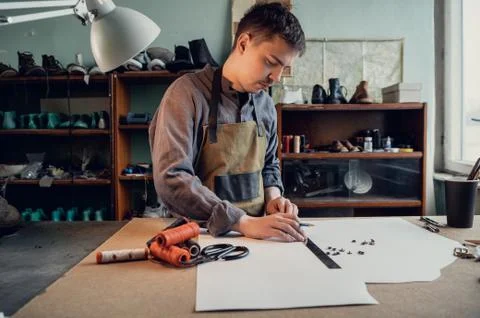 A young shoemaker makes a drawing for a pattern for leather shoes on a table in Stock Photos