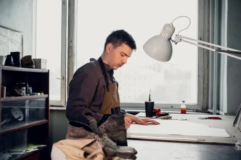 A young shoemaker makes a drawing for a pattern for leather shoes on a table in Stock Photos
