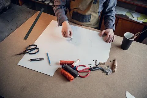 A young shoemaker makes a drawing for a pattern for leather shoes on a table in Stock Photos