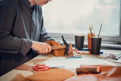 A young shoemaker using a hammer knocks down rivets on a leather product made Stock Photos