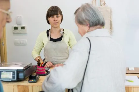 Young shopkeeper interacting with elderly lady at cash desk. Stock Photos