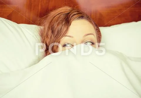 Young shy woman lying in bed hiding under duvet blanket Stock Photos