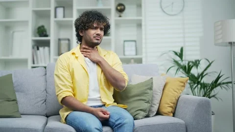 Young sick man coughs sitting at home on the couch holding hands to chest  Stock Footage 235505045