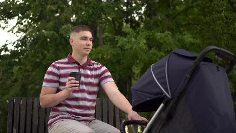 A young single father drinks coffee and rocks a stroller with a sleeping baby. A Stock Photos