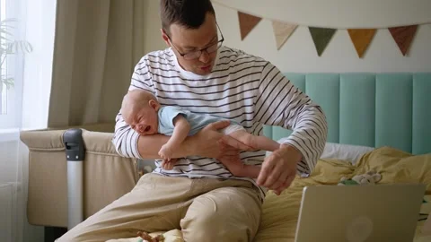 Young single father on paternity leave trying to work remotely on a laptop Stock Footage 344298985