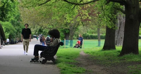 Young skater vaping on park bench while people stroll under spring trees Stock Footage 321019242