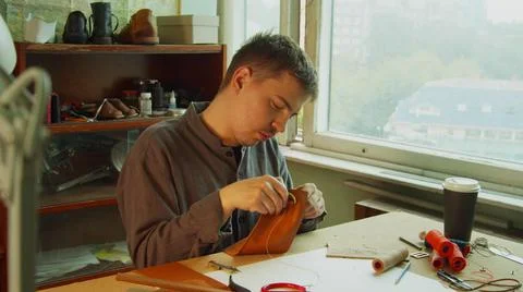 A young skilled tailor sews the element to a leather bag using a tailor's needle 스톡 사진