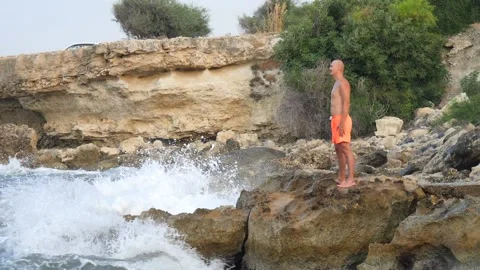 A young slender bald man in orange shorts and a naked torso stands on seashore 库存影片 134825396