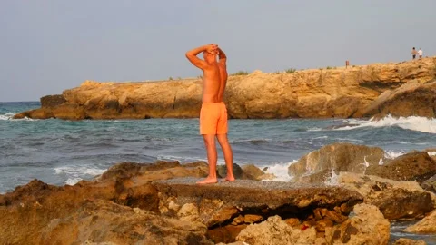 A young slender bald man in orange shorts and a naked torso stands 库存影片 134827622
