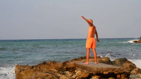A young slender bald man in orange shorts and a naked torso stands 库存影片 134830512