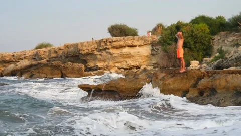 A young slender bald man in orange shorts and a naked torso stands on seashore Stock Footage 134836082