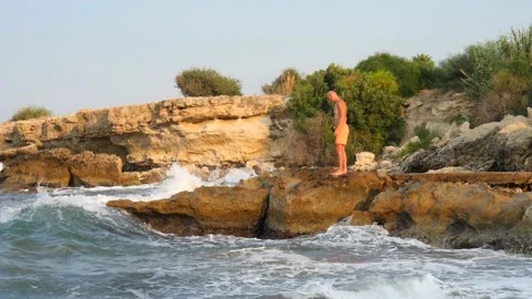 A young slender bald man in orange shorts and a naked torso stands on seashore Stock Footage 134840039