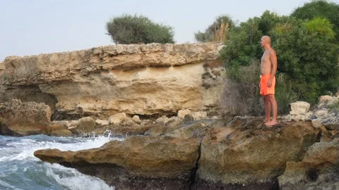A young slender bald man in orange shorts and a naked torso stands on seashore Stock Footage 134841559