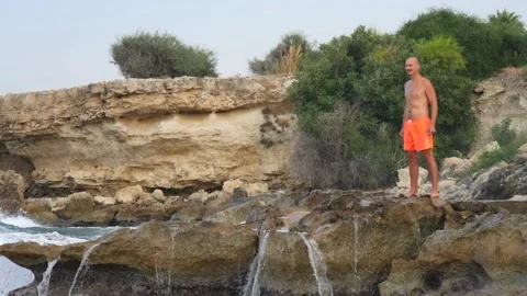 A young slender bald man in orange shorts and a naked torso stands on seashore Stock Footage 134842225