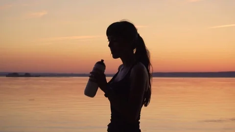 Young slender woman is drinking water from a bottle on the lake at sunset Stock Footage 77697341