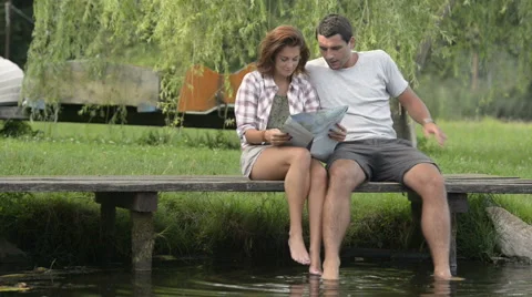 Young smiling couple look at map seated on lake jetty in summer day Vídeo Stock 64542708