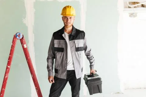 Young smiling engineer in work clothes and yellow hardhat holdin Stock Photos