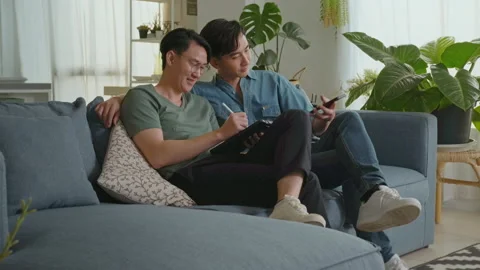 Young smiling gay couple using smartphone and relaxing in the living room at  Stock Footage 172287242
