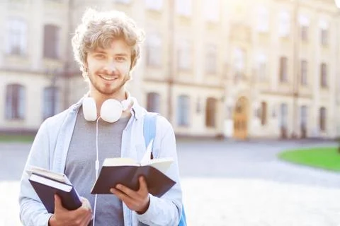 Young smiling man in backpack is holding books, standing outdoor on backgroun Stock Photos