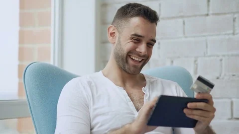 Young smiling man doing online shopping using digital tablet computer sitting at Stock Footage