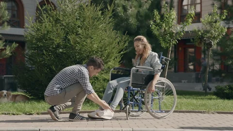 Young smiling man help pretty woman in a wheelchair tie shoelaces. Couple in Stock Footage 290916074