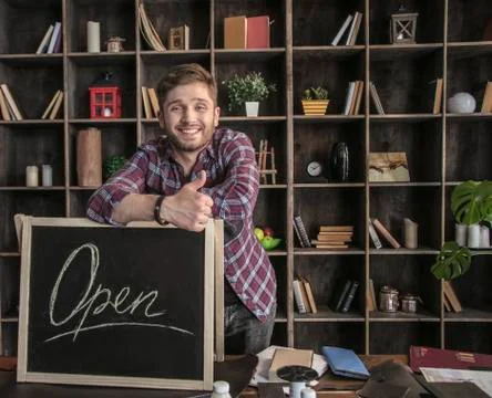 Young smiling man leather maker standing near open sign at wooden table Stock Photos