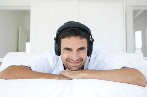 Young smiling man lying on bed, listening to music with headphones Stock Photos