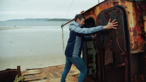 A young smiling man opens the door and goes inside of old rusty ship near beach Stock Footage 247285583