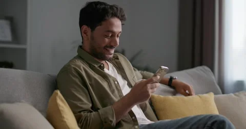 Young smiling man sitting on the sofa in living room, using app on smartphone. Video stock 248966745