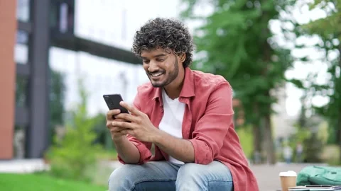 Young smiling man uses a mobile phone while sitting on a bench on street near  Stock Footage 302473085