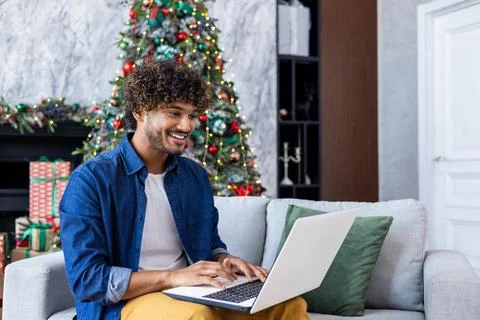 Young smiling man working remotely from home for Christmas sitting on couch in Stock Photos