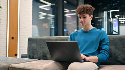 A young smiling man working using a laptop while sitting on a sofa in an office Stock Footage 235232442