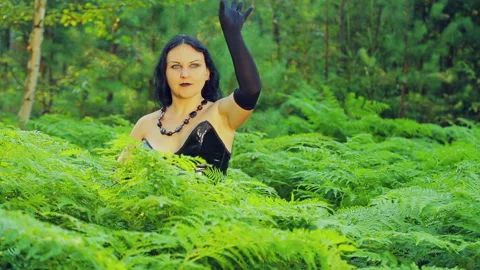 A young smiling witch in black clothes hides in a thicket of fern. Halloween Stock Footage 94459715