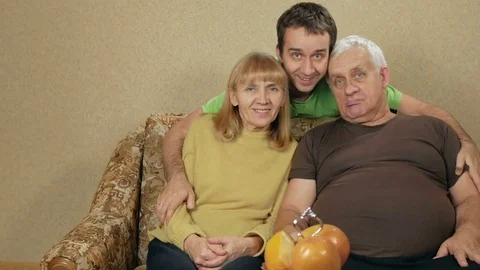 Young son of an elderly couple hugging at home on the couch. Everyone looks at Video stock 71246693