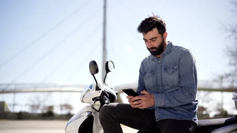 Young Spanish man using cell phone or smartphone sitting on his motor scooter Stock Footage 153251379
