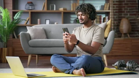 Young sportsman using mobile app on phone while sitting on mat on floor 스톡 동영상 334231007
