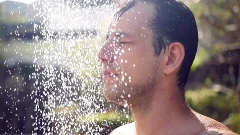 A young strong man stands under the shower in the sun, splashes of water Stock Footage 87448519