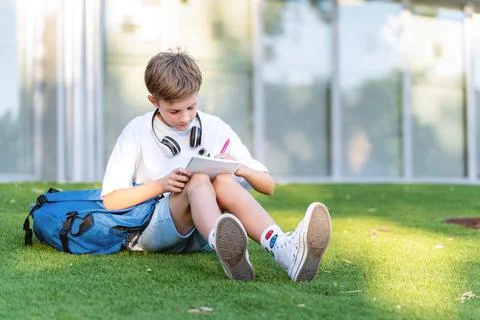Young student boy doing homework in notebook while sitting on the grass outdoors Foto stock