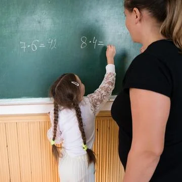 Young student calculates multiplication problem on chalkboard with teacher .. Stock Photos