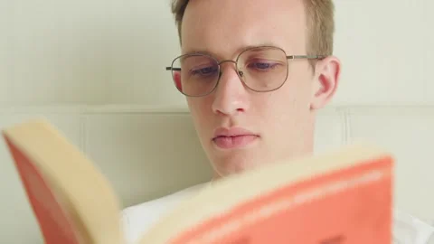 Young student in eyeglasses reading a paperback book indoors. Video stock 334848556