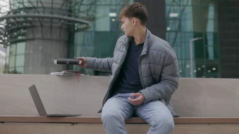 Young student reading a book on campus bench with laptop beside him Stock Footage 293762948