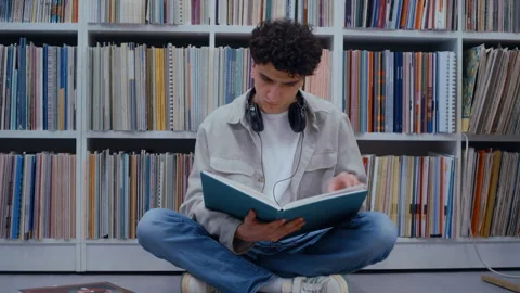 Young student reading a book sitting on the library floor 스톡 동영상 335124132