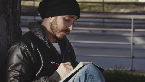 Young student sitting under a tree writing in a notebook 스톡 동영상 74729506