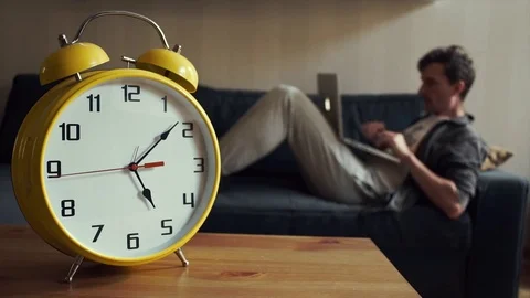 Young student using laptop sitting on couch at home. Big yellow clock is ticking Stock Footage 80814020