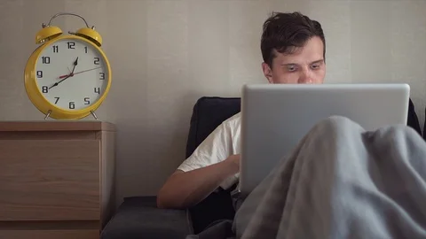 Young student using laptop sitting on couch at home. Big yellow clock is ticking Video stock 80815061