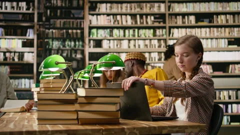 Young student working on laptop taking notes at library table with open book Stock Footage 337229194