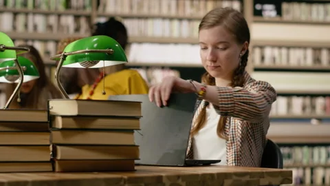 Young student working on laptop taking notes at library table with open book Stock Footage 337231930