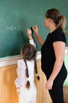 Young student works on multiplication problem on chalkboard with teacher in.. Stock Photos